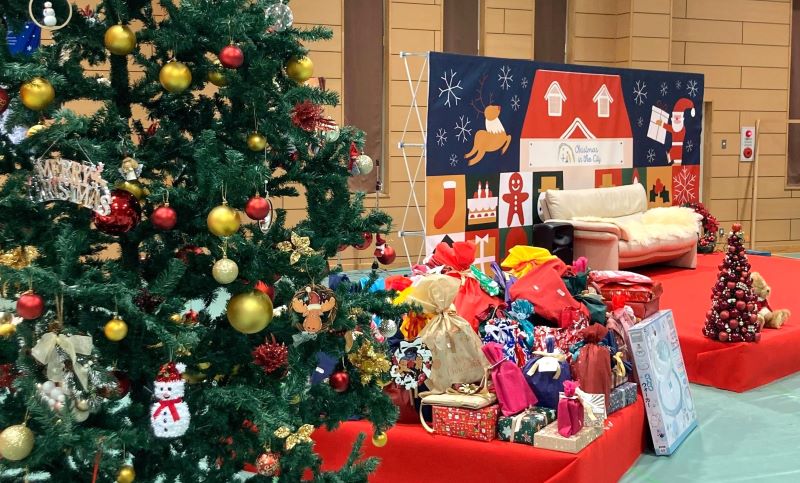 A Christmas tree, many wrapped presents, and a stage in the background with a Christmas-themed backdrop